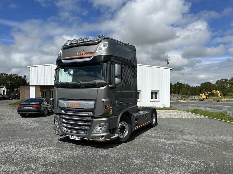 Scène extérieure d'un site de vente d'équipements de construction avec tracteur semi-remorque rouge et turquoise transportant une pelle sur roues