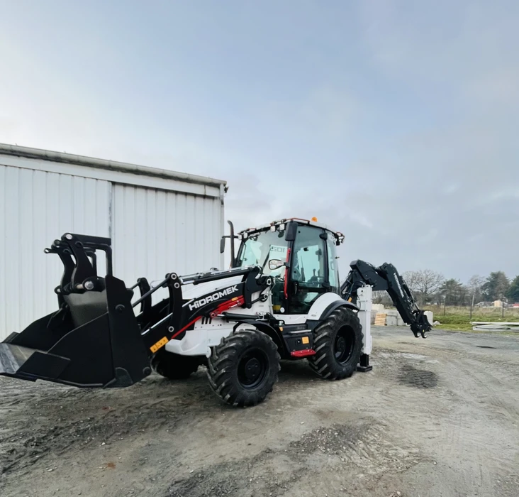 Scène extérieure d'un concessionnaire d'équipements de construction avec plusieurs engins de chantier stationnés devant un bâtiment métallique marqué "OZ TRUCK TRAILER"