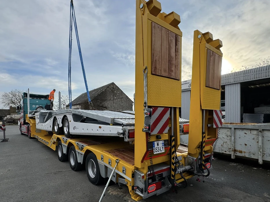 Scène extérieure d'un site de vente d'équipements de construction avec tracteur semi-remorque rouge et turquoise transportant une pelle sur roues